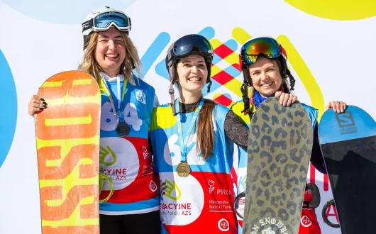 Trzy zawodniczki w strojach do snowbordu na tle baneru. Zdjęcie wykonane zimą na stoku.  