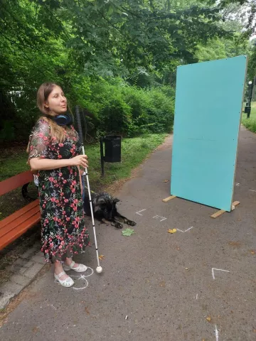 Kobieta z białą laską na tle zieleni i błękitnej tablicy odbijającej dźwięk 