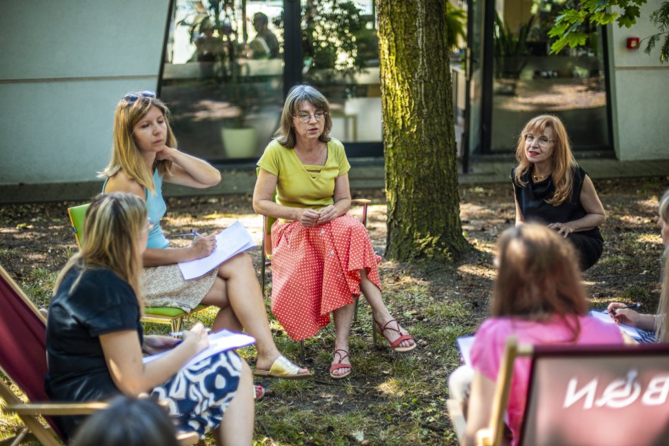 Grupa rozmawiających ze sobą osób siedzących w kręgu na krzesłach i leżakach wśród zieleni.  tyłu widoczny budynek BON. 